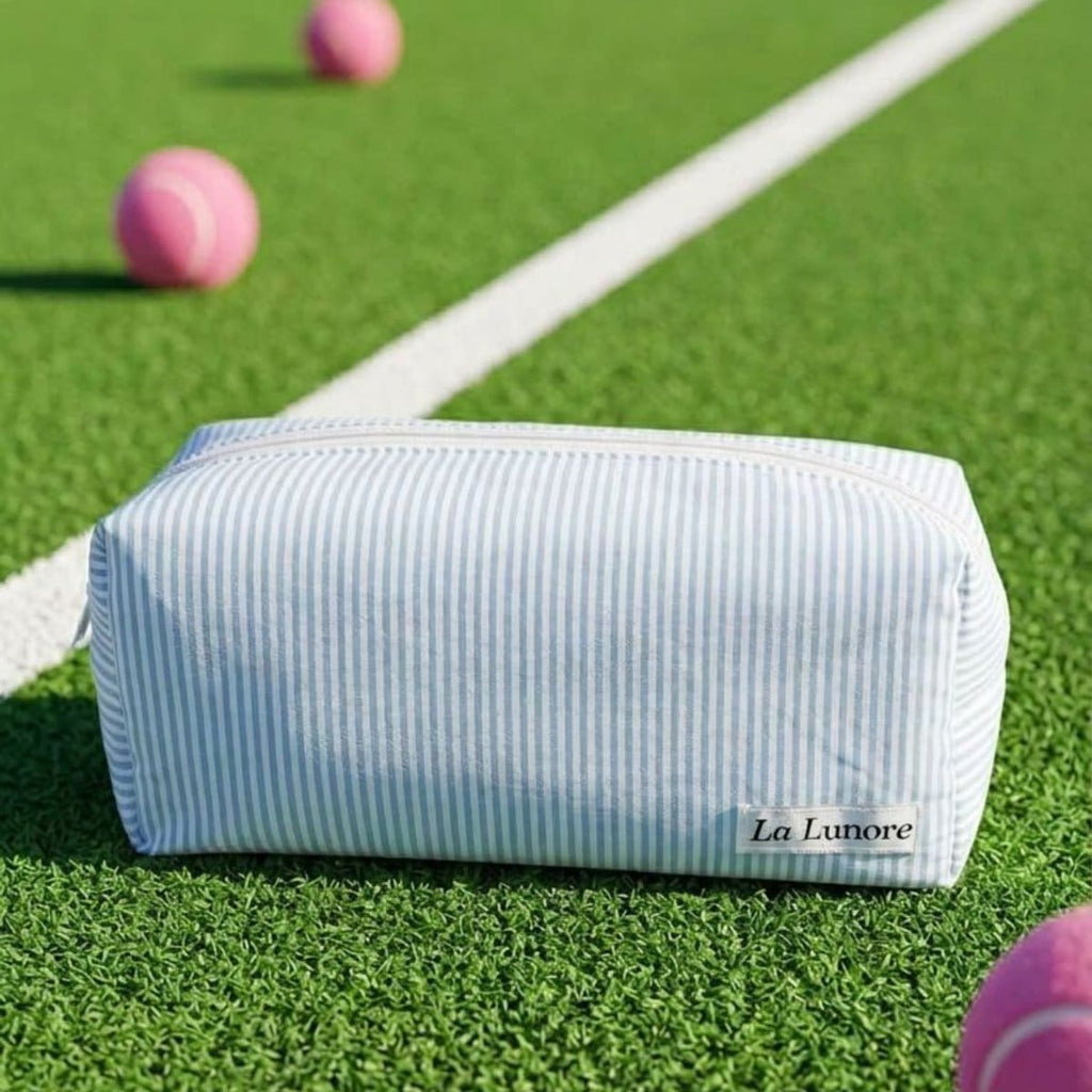Lichtblauw gestreepte La Lunore toilettas met fijne witte strepen op groen gras met roze tennisballen.
