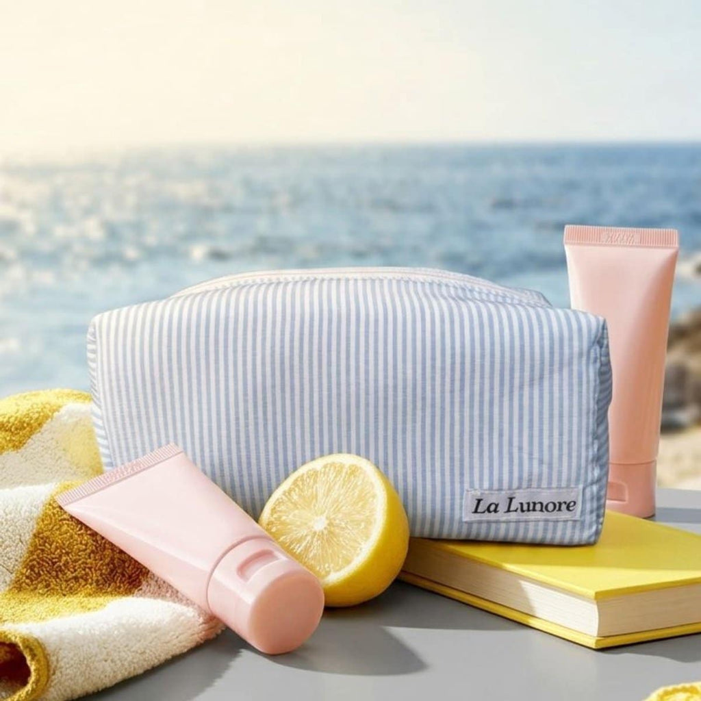 Lichtblauw gestreepte La Lunore toilettas naast een handdoek, citroen en twee roze tubes op een tafel aan zee.