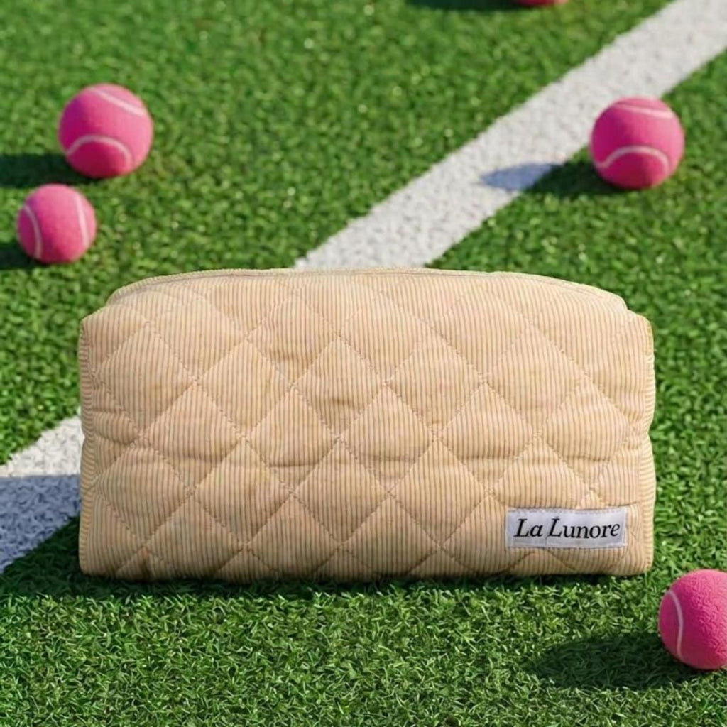 Crèmekleurige gewatteerde La Lunore toilettas met merklogo op groen kunstgras met roze tennisballen.