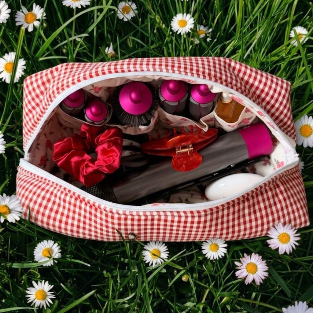 Open rood-wit geruite La Lunore Cherry toilettas met stylingtools, scrunchie en accessoires op kunstgras met madeliefjes.