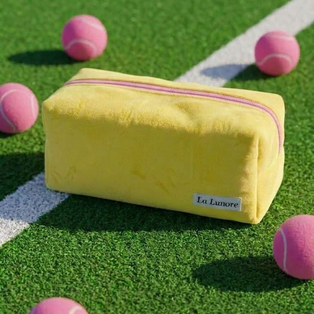 Gele La Lunore toilettas met roze rits op groen kunstgras met een witte lijn en roze tennisballen.